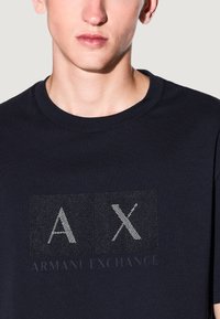 T-shirt bleu marine en coton, avec un logo texturé en fil argenté portant la mention "AX" et "ARMANI EXCHANGE" en dessous.