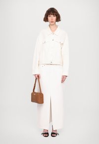 Weißjeansjacke mit Knöpfen, kombiniert mit einem weißen Maxirock mit Schlitz vorne. Braune Handtasche mit geprägtem Detail und schwarzen Sandalen.