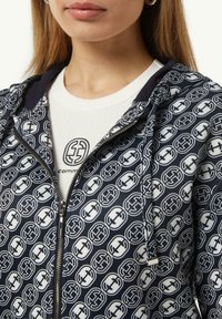 Marine blauwe hoodie met ritssluiting heeft een overal wit logo-patroon en een gladde textuur. Eronder is een witte crewneck t-shirt gelaagd.