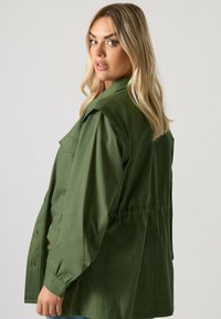 Vaaleahiuksinen nainen, jolla on löysät hiukset, pukeutuneena oliivinvihreään työtakkiin, katsoo yli olkansa neutraalia taustaa vasten.