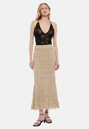 Donna con top nero in pizzo senza maniche e gonna lunga beige a fantasia, con tacchi neri con cinturini, in piedi davanti a uno sfondo semplice.