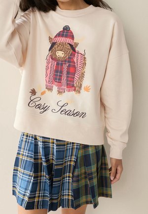 Sweat-shirt beige avec un taureau en dessin animé portant un chapeau et une écharpe à carreaux rouges et le texte "Cosy Season", associé à une jupe plissée à carreaux bleus et verts.