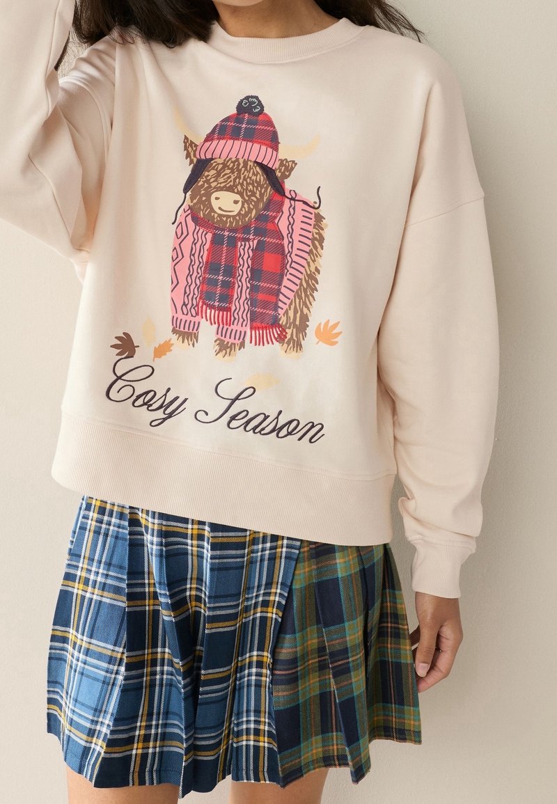 Sweat-shirt beige avec un taureau en dessin animé portant un chapeau et une écharpe à carreaux rouges et le texte "Cosy Season", associé à une jupe plissée à carreaux bleus et verts.