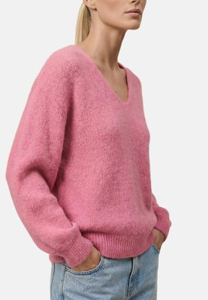 Femme portant un pull rose clair duveteux à col en V et un jean bleu clair, debout les mains dans les poches devant un fond uni.