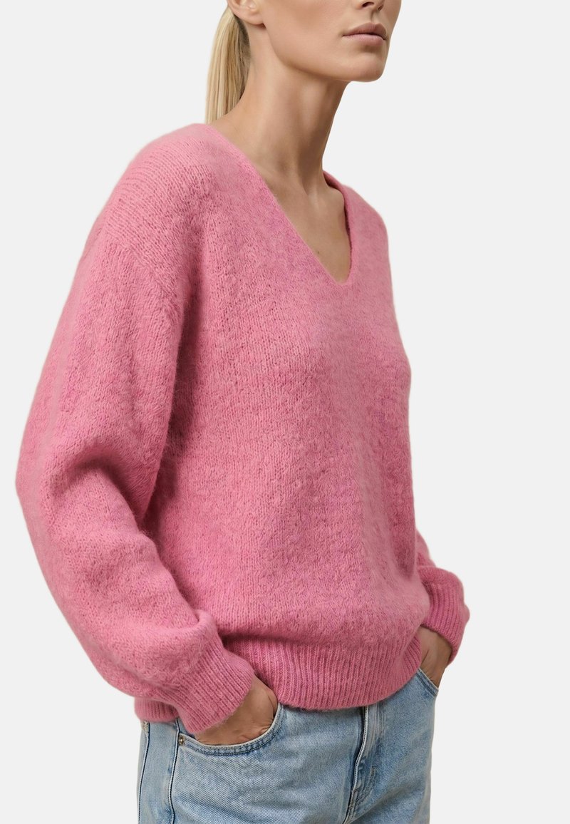 Femme portant un pull rose clair duveteux à col en V et un jean bleu clair, debout les mains dans les poches devant un fond uni.