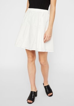 Vrouw draagt een witte rok met oogjespatronen, een zwarte mouwloze top en zwarte open teen met hak muilen, staand tegen een effen achtergrond.