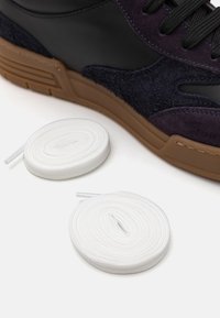 Kõrge sneaker mustast nahast ja tumesinises sametist, pruun kummist tallaga. Kaks valget ümmargust paela on asetatud lähedale.