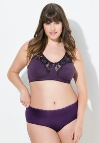 Kvinna med långt brunt hår iförd en lila bh med spetsdetaljer och matchande underkläder, poserande mot en vit vägg.