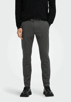 Mann trägt graue Slim-Fit-Hose, schwarzen Gürtel, schwarzes Langarmshirt und schwarze Schnürschuhe, steht mit einer Hand in der Tasche.