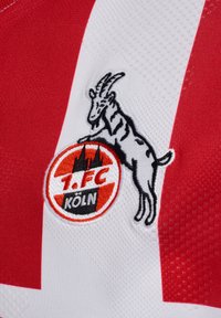 Rød og hvid fodboldtrøje med tekstureret stof. Har et broderet logo af 1.FC Köln med en ged og stiliserede tårne.