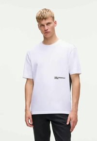 T-shirt de algodão branco de mangas curtas com o logótipo da Karl Lagerfeld Jeans no lado esquerdo; corte descontraído com um design simples. Calças pretas usadas por baixo.