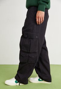 Person trägt lockere schwarze Cargo-Shorts mit geknöpften Taschen und weiße Sneaker mit grünen Swooshes auf einem hellgrünen Boden.