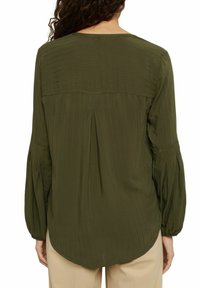 Blouse vert olive avec un design texturé et côtelé, encolure ronde et manches longues froncées ; présente un pli au centre du dos.