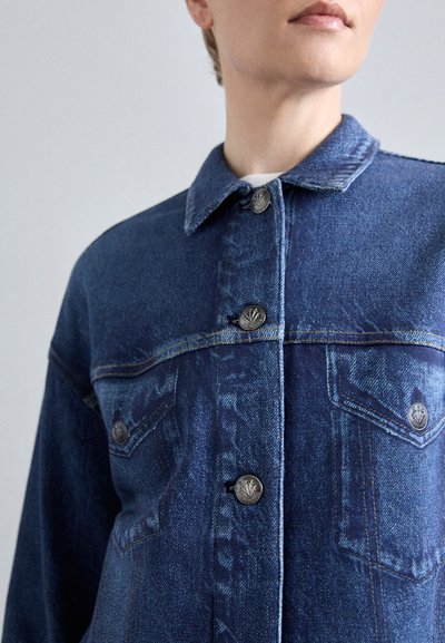 Jachetă din denim de culoare albastră închisă cu cusături contrastante; prezintă un guler clasic, două buzunare la piept și nasturi metalici.