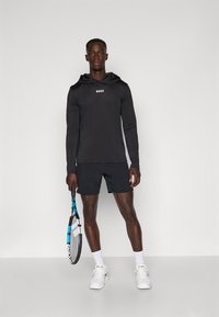 Mustapaitainen pitkähihainen huppu, jossa on "BOSS"-logo, yhdistettynä mustiin shortseihin. Malli pitää kädessään tennismailaa, ja hänellä on päällään valkoiset urheilukengät ja sukat.
