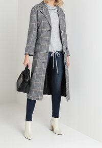 Vrouw met een lange geruite jas, grijze trui, donkere skinny jeans, witte enkellaarsjes en een zwarte handtas.