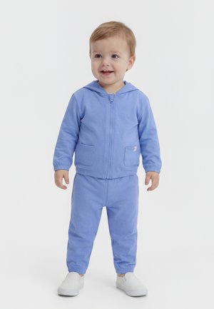 Νηπιάκια στέκονται χαμογελώντας, φορώντας ένα ανοιχτό μπλε μπουφάν με κουκούλα και φερμουάρ, ταιριαστό παντελόνι και λευκά παντοφλάκια, σε λευκό απλό φόντο.