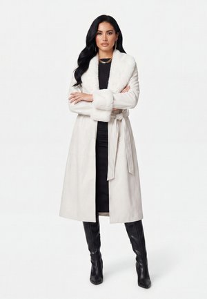 Donna con lunghi capelli neri indossa un cappotto bianco con colletto di pelliccia, un abito nero e stivali neri al ginocchio, in piedi con le braccia incrociate.