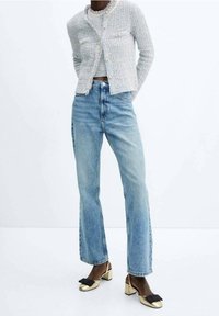 Cardigan gris clair texturé avec boutons en perles, associé à un jean bleu taille haute à jambes droites et des chaussures à talons blocs dorés avec des nœuds noirs.