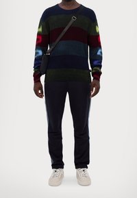 Mehrfarbig gestreifter Strickpullover mit langen Ärmeln, dunkle Hose mit hellen Seitenstreifen und weiße Sneakers. Eine schwarze Umhängetasche ist ebenfalls dabei.