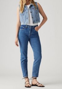 Ljusblå denimkort väst, vit linne, hög midja blå jeans och svarta sandaletter med remmar. Enkelt mönster med klassiska linjer.