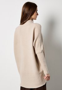 Suéter ribeteado beige con un cuello alto, corte oversize y detalles de botones en las mangas. Textura suave y diseño relajado.