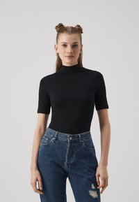 Zwarte body met korte mouwen en een hoge hals; gladde textuur. Gecombineerd met blauwe jeans met hoge taille en een versleten detail op de knie.
