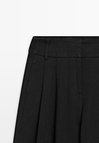Pantaloni neri plissettati con vita strutturata, tasche laterali e tessuto dalle texture, con un design pulito e minimalista.