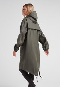 Chaqueta impermeable verde oliva con capucha, puños elásticos y un dobladillo trasero más largo. Cuenta con cordones ajustables en la cintura.