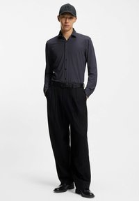 Camicia a maniche lunghe bianca e nera a pois, pantaloni neri plissettati, cintura nera e berretto nero con logo "HUGO" in rilievo. Scarpe nere completano il look.