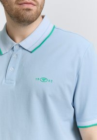 Ljusblå polo skjorta med en kontrasterande grön krage, tre knappar och en broderad logo med året "1962" på bröstet.