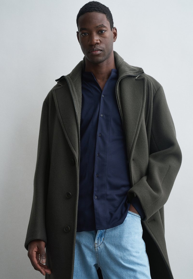 Oversized olijfgroene jas met een gestructureerde afwerking, grote kraag en knopen. Draag over een marineblauw shirt en lichtblauwe jeans.