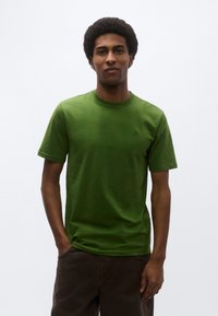 Πράσινο t-shirt με στρογγυλή λαιμόκοψη, κατασκευασμένο από βαμβάκι με κοντά μανίκια. Ομαλή υφή και διακριτική λεπτομέρεια λογ ότυπου στο στήθος. Συνδυάζεται με σκούρο καφέ παντελόνι.