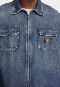 Giacca di denim blu, con zip frontale, due tasche sul petto con pattina e un'etichetta sulla tasca sinistra. Design del colletto classico.