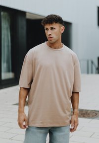 Světle béžové oversized tričko vyrobené z měkké látky, s klasickým kulatým výstřihem a krátkými rukávy, doplněné o světlé džíny.