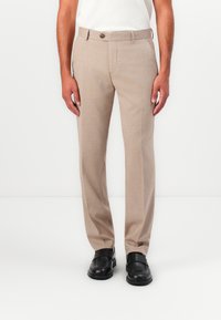 Beige szövetnadrág, texturált felülettel, egyenesen szabott fazonnal és egy gombos záródással, fekete slip-on cipővel párosítva.