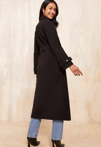 Melna garā jaka ar jostu un uzmavēm, apvienota ar gaiši ziliem džinsiem un melniem norādītajiem zābakiem. Gluda auduma, piegriezuma dizains.