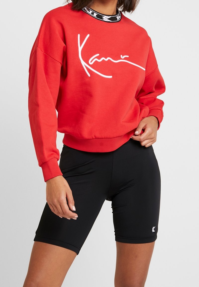 Personne portant un sweat-shirt rouge à manches longues avec un texte script blanc et un short ajusté noir sur un fond uni.