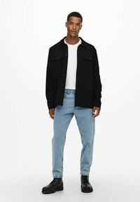 Camisa negra de botones con dos bolsillos en el pecho, llevada sobre una camiseta blanca, combinada con jeans de color azul claro y botas negras con cordones.