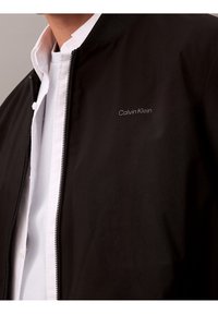 Schwarze Bomberjacke aus glattem, leichtem Material. Verfügt über einen Reißverschluss, einen gerippten Kragen und ein gesticktes "Calvin Klein"-Logo.