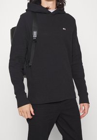 Sudadera con capucha negra de tela texturizada, puños acanalados y un pequeño logo en el pecho. Combinada con pantalones negros y una mochila.
