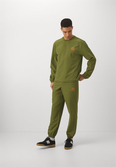 Olivgrüne Sweatshirt und Jogginghosen aus einem weichen Stoff, mit elastischen Bündchen und einem runden Logopatch. Die Schuhe sind schwarz mit Gummisohlen.