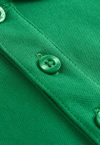 Žalias medvilnės polo marškinėlių detalė, kuri pasižymi tekstūruota medžiaga, mygtukų eilute ir prekės ženklu "Clarks", įspaustu ant mygtuko.