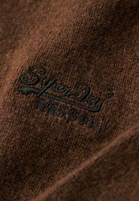 Tissu en laine marron avec une surface texturée, présentant un logo brodé "Superdry" en noir. Les détails montrent des points de couture et des fibres.