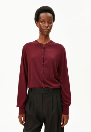 Frau mit kurzen Haaren, die einen bordeauxfarbenen, langärmeligen, geknöpften Pullover und schwarze, hoch taillierte Hosen vor einem einfarbigen Hintergrund trägt.