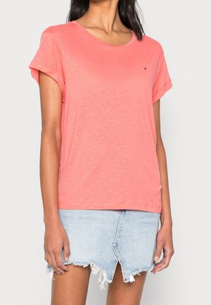 Kvinde iført koralrød T-shirt med korte ærmer og lyseblå denimnederdel med frynset kant, stående mod en ensfarvet baggrund.