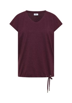 Frau lächelt, trägt eine pinke Kappe, ein weinrot gestreiftes Shirt, hellblaue Jeans, beigefarbene Sandalen und hält eine große schwarze geflochtene Tragetasche.