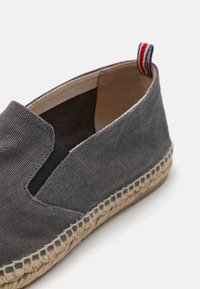 Chaussure slip-on en toile grise avec des inserts élastiques sur les côtés, semelle en corde de jute et un petit onglet rouge, blanc et bleu à l'arrière.