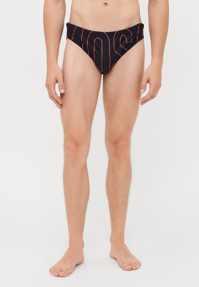 Uomo che indossa slip da bagno neri con scritte rosse, in piedi a piedi nudi su uno sfondo chiaro e semplice.