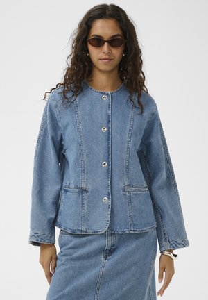 Kvinde med langt krøllet hår iført brune solbriller, en blå denimjakke med knapper og matchende denimskørt mod en ensfarvet baggrund.